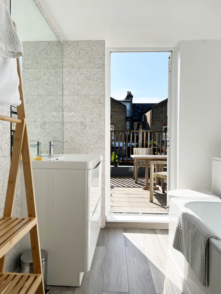 Bright bathroom with a white washing machine beside a tiled wall, a wooden ladder shelf, and a door opening to a sunny balcony with outdoor seating outside the door.