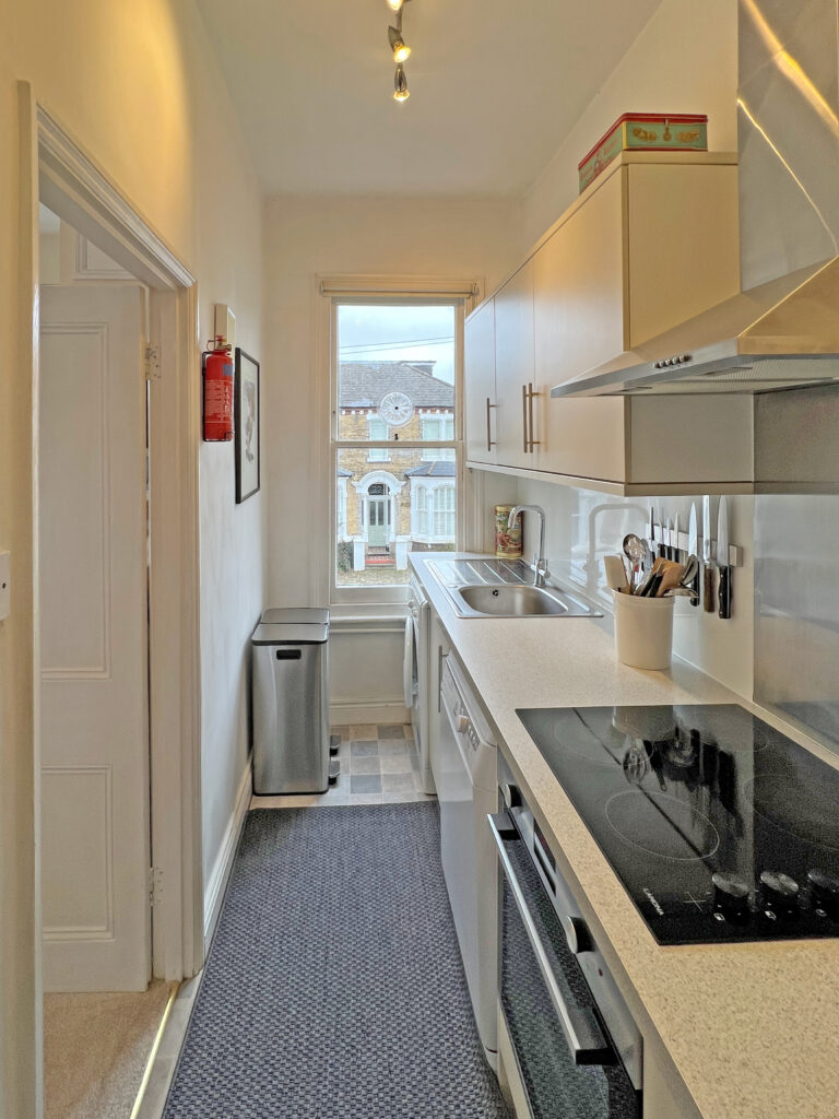 Narrow galley kitchen with white cabinets, sink and induction hob on the right, window at the far end.