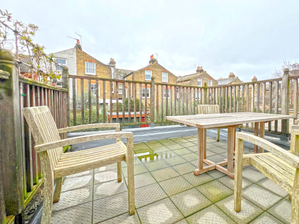 Wooden outdoor dining set on a rain-wet terrace with a railing, overlooking brick row houses under a cloudy sky