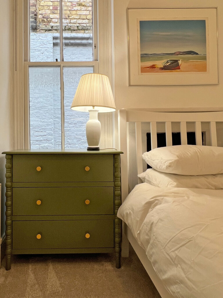 Green nightstand with a cream lamp beside a white bed, framed beach artwork on the wall, and a window showing a brick exterior outside the room edge.